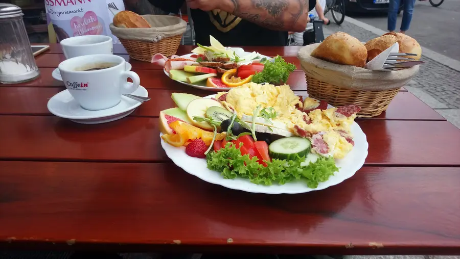 Frühstück draußen auf dem Holztisch: Kaffee, Brötchenkorb und ein Teller mit Rührei, Obst und Gemüse