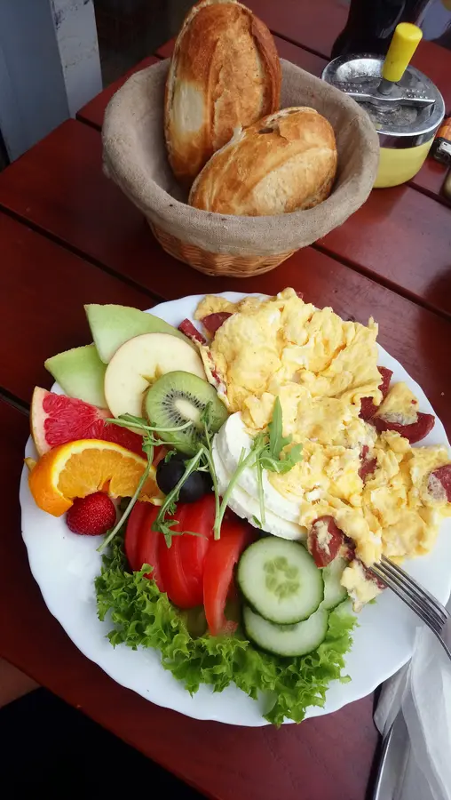 Rührei-Frühstück mit Obst, Gemüse und Frischkäse, dahinter ein Korb mit Schrippen auf dem roten Holztisch